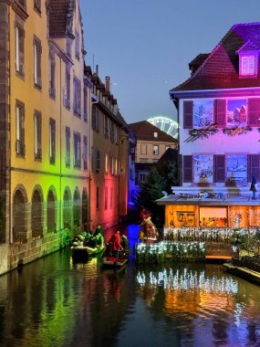 Colmar, Fransa 16 Aralık 2024: Colmar 'da ışıklandırılmış binalar, Noel süslemeleri, kanalda kayıklar ve arka planda parlayan dönme dolap ile birlikte suda yansımalar olan renkli bir gece sahnesi.