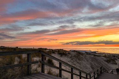 Kum tepeleri ve deniz üzerinde canlı günbatımı, kıyı şeridinin üzerinde parlayan pastel bulutlar. Tahta merdivenler sahile doğru sakin bir sahile çıkıyor. Yumuşak renkler ve geniş kompozisyon onu bir masaüstü arkaplanı veya duvar kağıdı olarak ideal kılar.