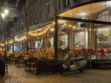 Bruges, Belçika, 25 Aralık 2025. Bruges 'de geceleri yılbaşı ışıklarıyla süslenmiş rahat restoran terası. Sıcak şenlik ışıltısı, boş sandalyeler ve park etmiş bir scooter tarihi kasabada kış akşamlarının cazibesini yaratır..