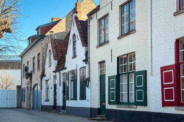 Bruges, Belçika, 26 Aralık 2025. Beyaz evleri ve renkli panjurları olan sessiz bir kaldırım taşı caddesi. Güzel gablonlar, rahat pencereler ve kış güneşi tarihi cazibenin tatlı bir kartpostal görüntüsünü oluşturur..