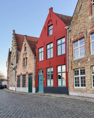 Bruges, Belçika, 26 Aralık 2025. Bruges 'de sakin bir kaldırım taşı caddesindeki renkli Flamanların basamaklı evleri yakından görülüyor. Turkuaz kapılı ve pencereli parlak kırmızı bir cephe tarihi eski kasabadaki geleneksel tuğla binaların arasında duruyor.