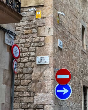 Barselona, İspanya 2 Ekim 2025: Ortaçağ taş cephesinde trafik kısıtlamaları, güvenlik kameraları ve sokak isimlerinin yer aldığı tabela kümesi. Küçük bir dini duvar figürü Barcelonas eski şehir mimarisinin tarihi karakterini vurguluyor