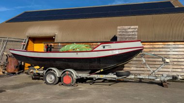 Açık motorlu balıkçı teknesi, çatısı güneş panelleri olan ahşap sanayi binasının yanında asfalt üzerinde duran metal bir tekne römorkuna monte edilmiş. Deniz taşımacılığı, tekne deposu, sürdürülebilir enerji ve eğlence amaçlı tekne konsepti.