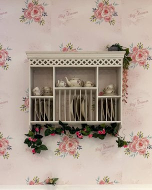 Zoetermeer, Hollanda 2 Mart 2026: Eskitilmiş çay setiyle dekoratif duvar rafı, çiçekler üzerine yerleştirilmiş tabaklar ve fincanlar, yumuşak pastel renklere sahip retro tarzı bir kafe içinde güller.