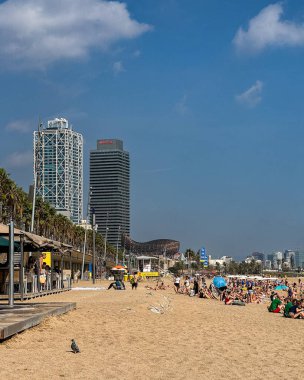 Barselona, İspanya - 4 Ekim 2025: Barceloneta sahilinde güneşli havanın tadını çıkaran turistler ve yerliler, ünlü Barcelona gökdelenleri arasında Hotel Arts, Mapfre Tower ve Port Olimpic bölgesi de bulunuyor..