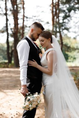 Bir çift düğün törenlerinde ormanda bir anı paylaşıyor. Damat takım elbise giyiyor ve gelin bir buket tutuyor. Güneş ışığı ağaçların arasından süzülür.