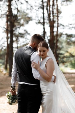 Gelin ve damat ormanlık alanda yakın dururlar. Damat, gelini aşağı bakarken kucağına alır. Güneş ışığı arka plandaki ağaçların arasından süzülerek doğal bir ortam yaratır..