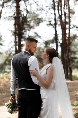 Bir çift, orman ortamında düğün kıyafetleri giyerek hassas bir anı paylaşıyor. Birbirlerine bakıyorlar, gün boyunca ağaçların altında birlikte vakit geçiriyorlar..