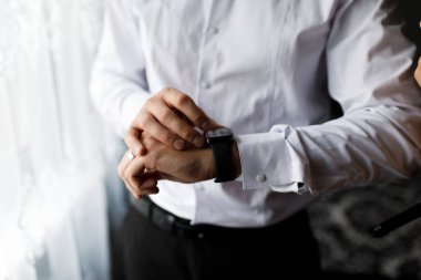 Bir adam gömleğinin manşetini ayarlıyor ve sabahleyin kapalı bir toplantı sırasında pencerenin yanındaki saatine bakıyor. Zaman ve görünüme odaklanıyor..