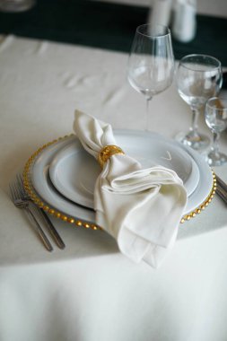 The table is arranged with a white tablecloth. A large plate sits under a smaller plate, both set with a folded napkin. Glasses filled with water are placed beside the plates.