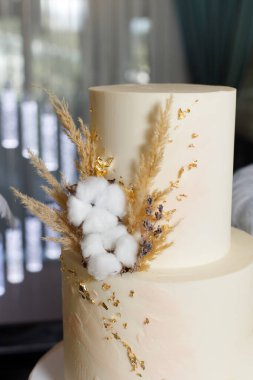 Bir pasta kapalı bir masada duruyor. Düz bir yüzeyi vardır ve pamuk, kurutulmuş çiçekler ve altın aksanlarla süslenmiştir. Yumuşak ışık arkaplanda filtrelenir.