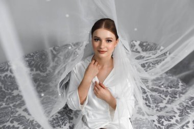 Beyaz bir bornoz giymiş bir gelin kanepede oturuyor. Saçlarını düzeltiyor ve etrafındaki perdeler gibi gülümsüyor. Yumuşak ışık odayı dolduruyor..