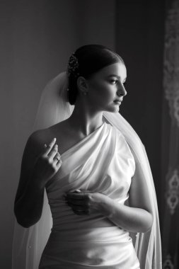 Gelin bir pencerenin yanında durarak düğün gününe hazırlanıyor. Uzağa dikkatlice bakarken bir elbise ve peçe takıyor. Işık kutlamadan önceki anı güçlendiriyor..