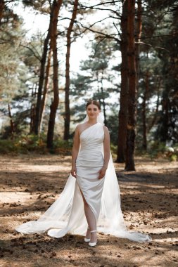 Bir gelin ormanda poz verir. Beyaz bir elbise giyiyor ve ağaçların arasında çıplak zeminde duruyor. Güneş ışığı dalların arasından süzülüp şu an için doğal bir zemin oluşturuyor..