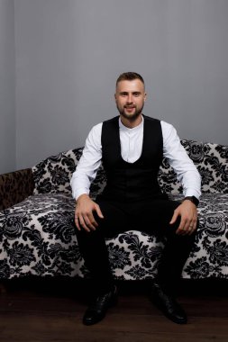 Resmi kıyafetler giymiş bir adam kanepede oturur. Onun kısa saçı ve sakalı var. Odada gri duvarlar ve desenli bir kanepe var. Poz verirken kendinden emin ve rahat görünüyor..