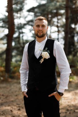 Bir adam ormanda siyah yelek ve beyaz gömlek giyerek poz veriyor. Göğsüne takılı bir çiçek var ve bir olaya hazır, uzun ağaçların arasında güvenle duruyor..
