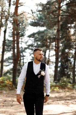 Bir adam yelek ve gömlek giymiş, göğsünde bir çiçekle ormanda yürüyor. Güneş ağaçların arasından parlıyor, parlak ve doğal bir ortam yaratıyor..