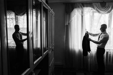 Bir adam büyük pencereli bir odada duruyor ve dolaptan takım elbise ceketi çıkarıyor. Sabah ışığında bir olaya hazırlanırken yansıması camda beliriyor..