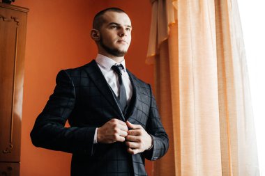 Turuncu duvarlı bir odanın penceresinin yanında bir adam duruyor. O bir takım elbise giyiyor ve ceketini ayarlıyor. Güneş ışığı perdelerin arasından gelir ve parlak bir atmosfer yaratır..