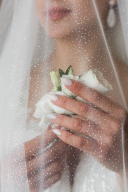 Gelinlik giymiş bir kadın göğsünde beyaz güller tutar. O, yağmur yağdığını öne süren su damlacıklarıyla bir peçenin arkasında. O an neşeyi ve duyguyu yakalar..