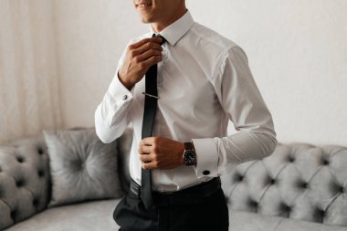 Bir adam resmi bir etkinliğe hazırlanıyor. Oturma odasında dururken kravatını düzeltiyor. Arka plandaki kanepe sahneye modern bir dokunuş katıyor..