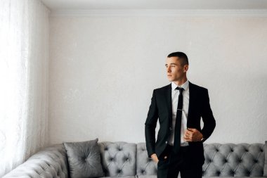 Oturma odasındaki pencerenin yanında takım elbiseli bir adam duruyor. Perdelerden süzülen ışıkla kravatını tutarken düşünceli görünüyor. Odada gri bir kanepe ve beyaz duvarlar var..