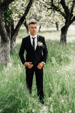 Genç bir adam yeşil bir alanda duruyor. O resmi bir takım elbise giyiyor ve ceketinde bir çiçek var. Arka planda uzun ağaçlar var. Gündüz oldu ve güneş parlıyor..