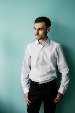 Mavi bir duvarın yanında genç bir adam duruyor. O, beyaz bir gömlek ve siyah pantolon giyiyor. Yan tarafa bakıyor ve gün boyunca derin düşüncelere dalmış gibi görünüyor..