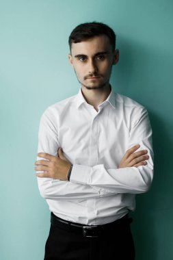 Açık renk gömlekli genç bir adam kollarını koyu renkli bir duvarın önünde kavuşturur. Kapalı mekanda ciddi bir ifadeyle doğrudan izleyiciye bakıyor..