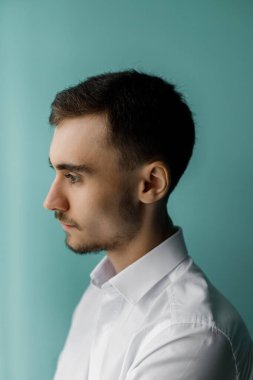 Bir adam turkuaz bir duvarın yanında duruyor. Beyaz bir gömlek giyer ve dik dik bakar. Yumuşak ışık yandan geliyor, onun özelliklerini net bir şekilde gösteriyor..