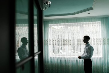 Bir adam pencerenin yanındaki parlak bir odada tek başına duruyor. Bardaktaki yansımasına bakıyor. Perdeler güneş ışığını filtreler ve etrafında yumuşak bir parıltı yaratır..