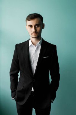 Genç adam elleri ceplerinde duruyor siyah takım elbise ve beyaz gömlek giyiyor. O, açık mavi bir duvara karşı kameraya bakarken ciddi görünüyor..