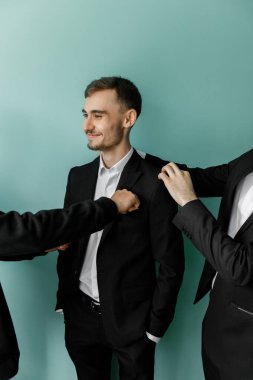 Üç kişi, takım elbiseli bir adama, sağlam bir geçmişi olan bir odada kıyafetlerini ayarlayarak yardım ediyor. Sosyal bir toplantıya hazırlanırken odaklanmış ve meşgul görünüyorlar..