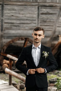 Siyah takım elbiseli ve beyaz gömlekli bir adam dışarıda ceketini düzeltiyor. Sahnede arka planda ahşap elementler ve yeşillik yer alıyor..