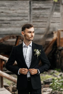 Damat düğününe hazırlanırken takım elbisesini ayarlıyor. Ahşap bir yapının yanında duruyor, düşünceli ve tören için hazır görünüyor. Açık hava ayarı anı artırıyor..