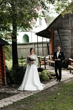 Beyaz elbiseli bir gelin, ahşap yapıları olan bir bahçede damadına yaklaşıyor. Çift yeşilliğin ortasında duruyor, düğün töreni için hazırlar..