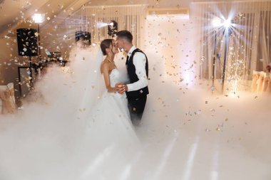 Yeni evli bir çift, dönen sis ve düşen konfeti arasında yakından dans ederek neşe ve kutlamalarla dolu dekore edilmiş bir düğün mekanında büyülü bir atmosfer yaratıyor..