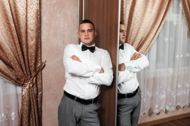 Sivri bir smokin giymiş genç bir adam iyi dekore edilmiş bir odada güven içinde duruyor. Kolları çapraz bir şekilde aynanın önünde duruyor. Kıyafetini ve davranışlarını gösteriyor..