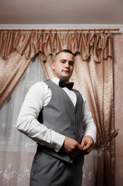 Gri takım elbiseli ve papyonlu genç bir adam şık bir odada duruyor, özel bir olaya hazırlanıyor, atmosferde kendine güven ve zarafet sergiliyor..