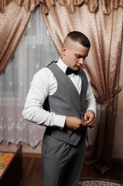 Damat düğününe hazırlanırken takım elbisesini ayarlıyor. Ayar, yumuşak ışıklandırma, zarif perdeler ve arka planda sıcak bir atmosfer yaratan eski tarz bir tablo içeriyor..