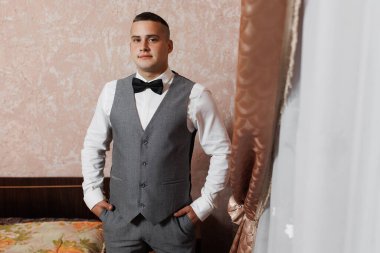 İyi giyimli genç bir adam şık bir odada duruyor, gri bir takım elbise ve papyon takıyor. Zarif ayar, resmi bir olay için uygun çiçek dekoru içerir.