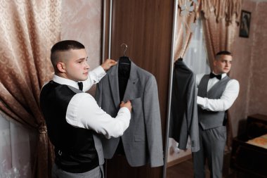 Resmi giyinmiş genç bir adam bir aynanın önünde dururken ceketini ayarlar. Samimi odada zarif perdeler ve düğünden önce neşeli bir atmosfer var..