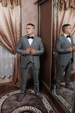 İyi giyimli genç bir adam, gri bir takım elbise ve papyon giymiş, lüks bir iç dekorasyon sergileyen süslü bir aynanın önünde poz verir..