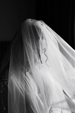 Bir gelin, uzun peçesiyle yüzünü örten bir odada duruyor. Odaklanmış ve tören için hazır görünüyor. Işık pencereden girer, yumuşak gölgeler yaratır..