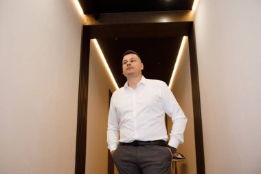 Beyaz gömlekli ve gri pantolonlu bir adam modern bir koridorda güvenle yürüyor. Koridor, duvarlardaki sıcak ışıkla aydınlanıyor..