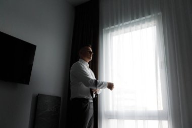 Bir adam otel odasında dikiliyor, perdeleri açık bir pencerenin yanında kravatını düzeltiyor. Kendini bir olaya ya da sabah güneşinde buluşmaya hazırlar..