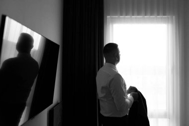 Genç bir adam sabah saatlerinde bir otel odasında duruyor. Elinde bir ceket tutarken pencereden dışarı bakıyor. Duvara monte edilmiş ekranda onun bir yansıması görünüyor..