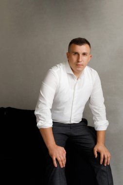 Bir adam elleri dizlerinin üzerinde bir sandalyeye oturur. Beyaz gömlek ve siyah pantolon giyiyor. Arka plan hafif, ve odak noktası onun duruşu ve ifadesi..