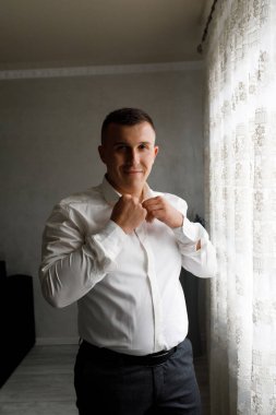 Adam pencerede gömleğini düzelterek bir olaya hazırlanır. Yumuşak ışık odaya girer, ortamın altını çizer. Görünüşüne odaklanmış görünüyor..