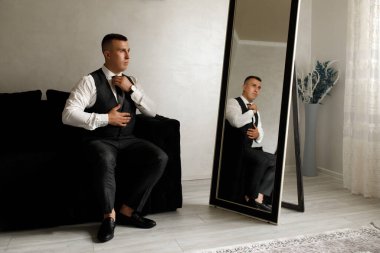 Adam siyah bir kanepede oturuyor, kravatını düzeltiyor ve aynada kendini seyrediyor. Güneş ışığı pencereden içeri girer ve odada parlak bir atmosfer yaratır..